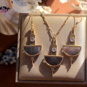 Pode incluir: Um colar de ouro e brincos combinando com um design geom&eacute;trico. O pingente e os brincos apresentam um semic&iacute;rculo de madrep&eacute;rola iridescente com uma borda dourada e um strass transparente no centro.