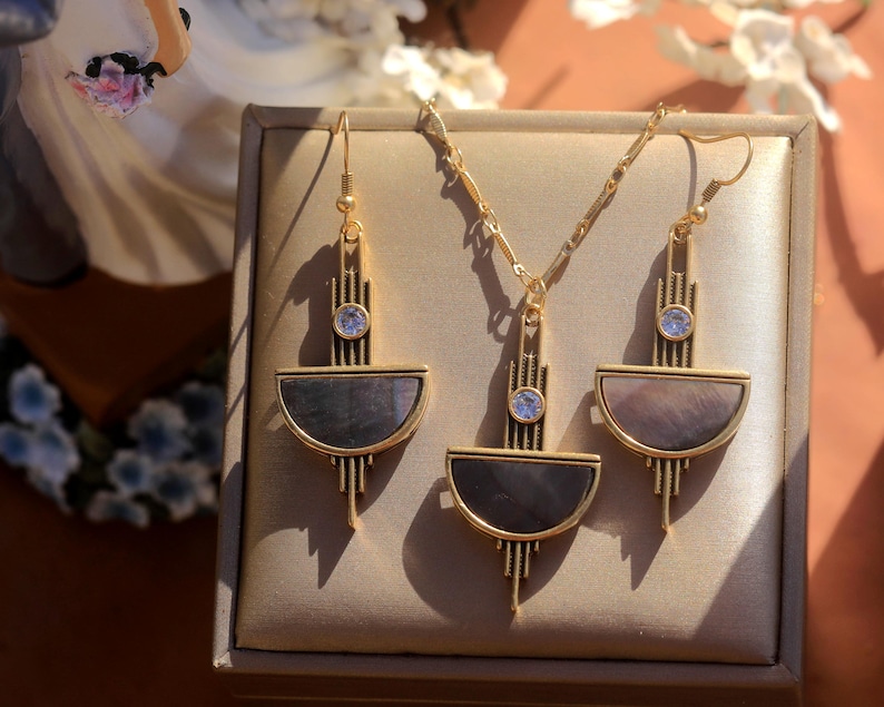 Pode incluir: Um colar de ouro e brincos combinando com um design geom&eacute;trico. O pingente e os brincos apresentam um semic&iacute;rculo de madrep&eacute;rola iridescente com uma borda dourada e um strass transparente no centro.