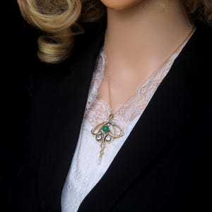 May include: A gold-toned pendant necklace featuring a central green gemstone, surrounded by clear crystals and a dangling pearl. The necklace is worn over a white lace top and a black blazer.