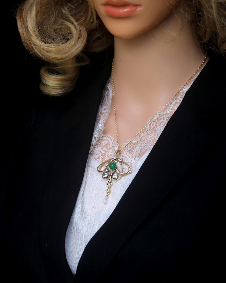May include: A gold-toned pendant necklace featuring a central green gemstone, surrounded by clear crystals and a dangling pearl. The necklace is worn over a white lace top and a black blazer.