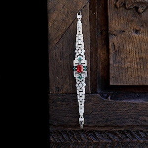 Peut inclure: Un bracelet en argent orné d'une pierre rouge centrale, entourée de petites pierres vertes et transparentes. Le bracelet présente un design Art Déco, avec des formes géométriques et un motif symétrique. Il est présenté sur une surface en bois sombre.