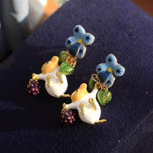 May include: A pair of earrings featuring a white mouse holding a purple berry and a blue flower with green leaves.