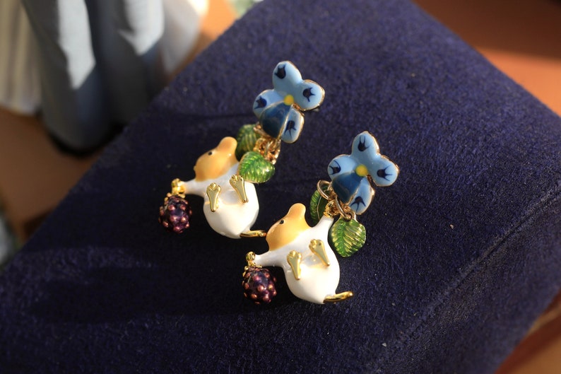 May include: A pair of earrings featuring a white mouse holding a purple berry and a blue flower with green leaves.