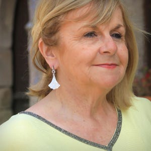 May include: A woman wearing a light yellow top with a beaded neckline and a pair of white, fan-shaped earrings. The earrings are attached to a small silver hoop. The woman has blonde hair.