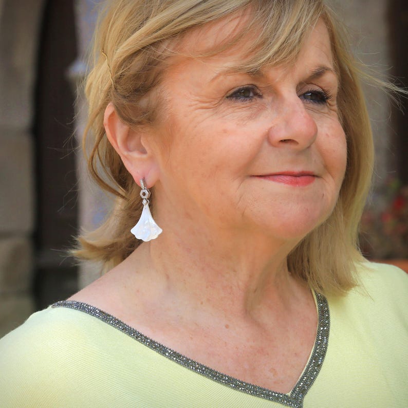 May include: A woman wearing a light yellow top with a beaded neckline and a pair of white, fan-shaped earrings. The earrings are attached to a small silver hoop. The woman has blonde hair.