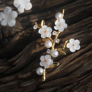 May include: A gold-tone brooch with a delicate floral design. The brooch features white flowers with pearl accents and clear rhinestones.