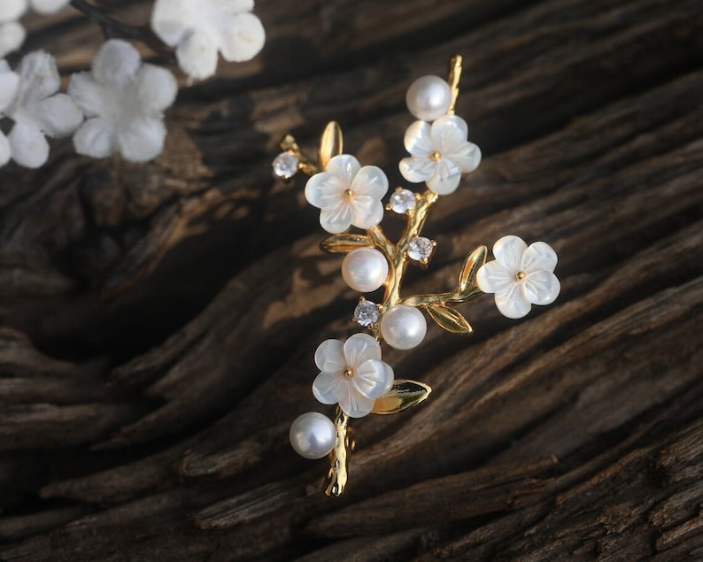 May include: A gold-tone brooch with a delicate floral design. The brooch features white flowers with pearl accents and clear rhinestones.