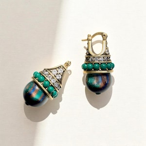 Puede incluir: Un par de pendientes de aro dorados con un colgante en forma de bellota. El colgante incluye una cuenta iridiscente de color verde oscuro y bronce, una fila de pequeñas esferas de color turquesa y una banda de piedras claras engastadas en oro.