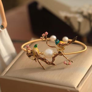 May include: A gold bangle bracelet with a delicate branch design featuring two colorful birds and a pearl. The birds are yellow, green, and blue with black accents. The branch is adorned with pink blossoms.
