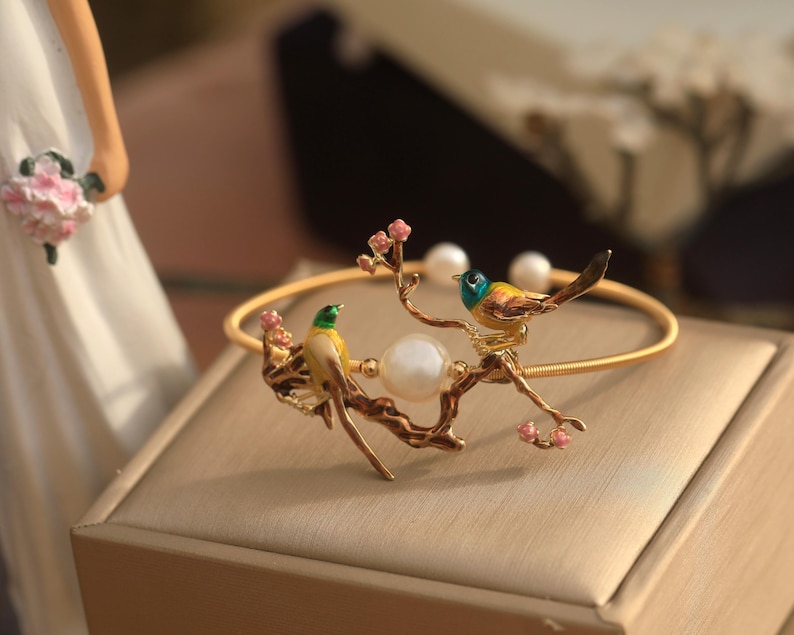 May include: A gold bangle bracelet with a delicate branch design featuring two colorful birds and a pearl. The birds are yellow, green, and blue with black accents. The branch is adorned with pink blossoms.