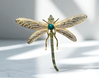 May include: A gold dragonfly brooch with crystal-studded wings and a teal central stone. The body and tail are embellished with green gemstones. This elegant brooch, with its detailed design, would make a striking addition to any outfit.