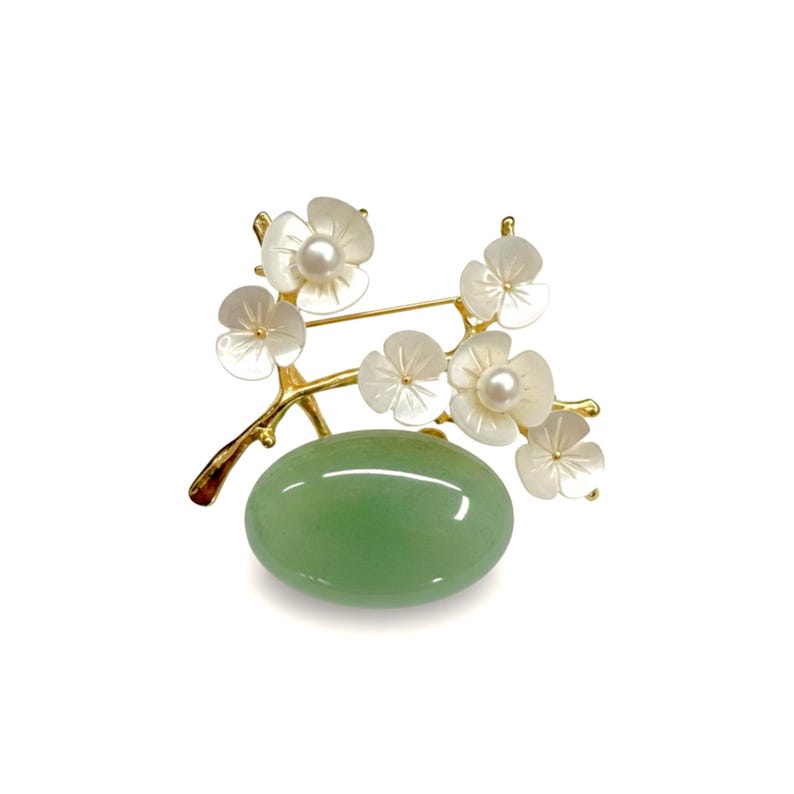May include: A gold-toned brooch featuring a branch with white flower blossoms and a central oval-shaped green stone. Each flower has a pearl center. The brooch is set against a white background.