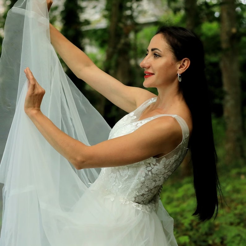 May include: A woman in a white lace wedding dress with a tulle skirt holds the veil up with one hand. She is standing in a forest with green foliage.
