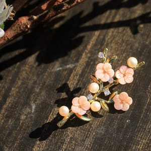 May include: A gold-tone brooch with a delicate floral design. The brooch features pink flowers with white centers and pearl accents.