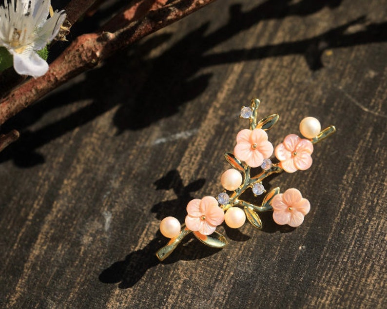 May include: A gold-tone brooch with a delicate floral design. The brooch features pink flowers with white centers and pearl accents.