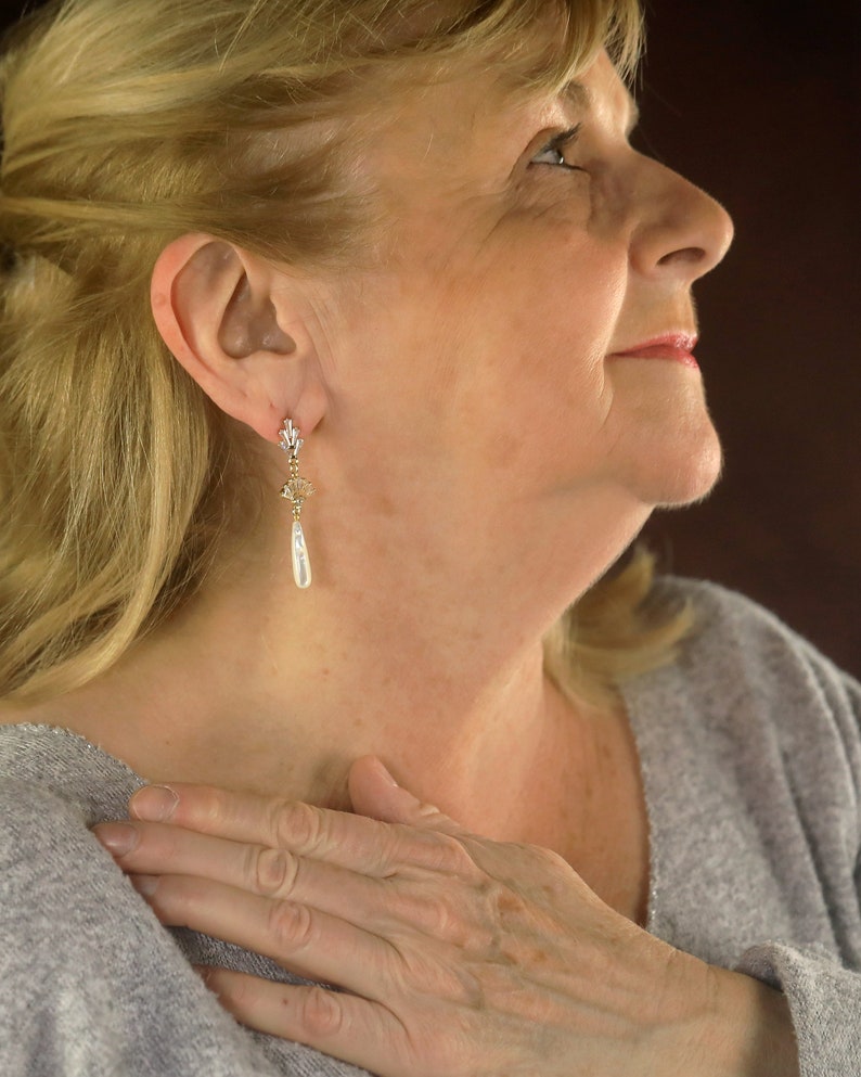 Op de afbeelding: Een vrouw met blond haar draagt een grijze trui en gouden oorbellen met een parel hanger.