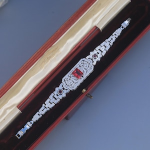 Puede incluir: Un brazalete de estilo Art Déco con una gema roja rectangular en el centro y gemas rojas más pequeñas en cada extremo. El brazalete es plateado y está engastado con numerosas gemas pequeñas y transparentes. Descansa dentro de un joyero rojo.