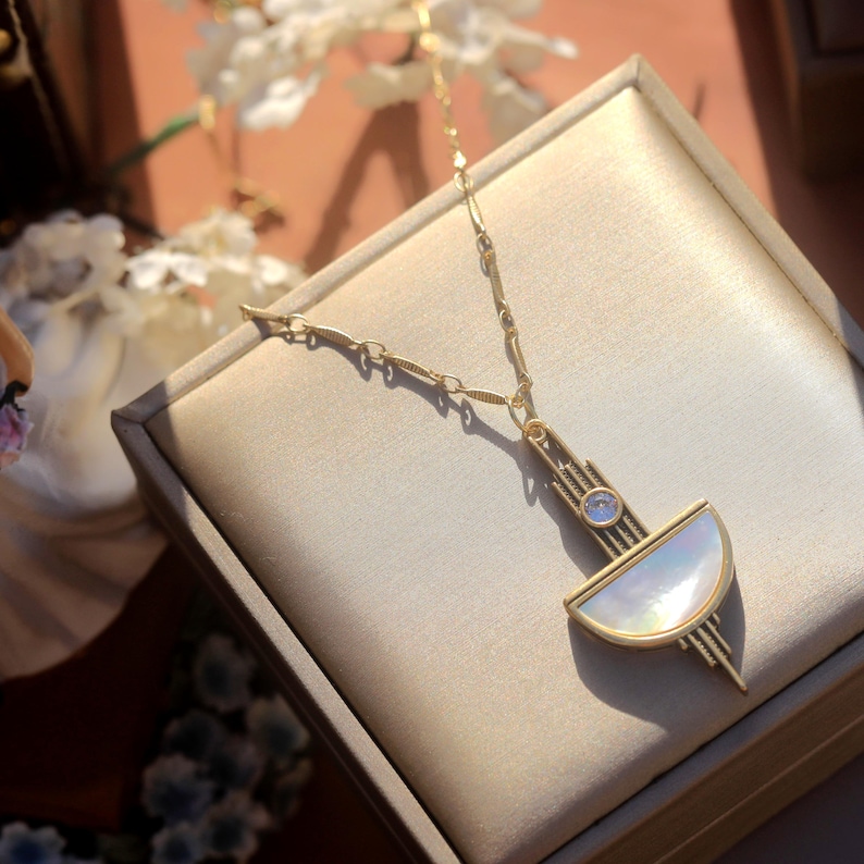 May include: A gold-toned necklace featuring a geometric pendant with a semi-circular iridescent mother-of-pearl inlay and a small round clear stone. The pendant is suspended from a delicate gold-toned chain with textured oval links.