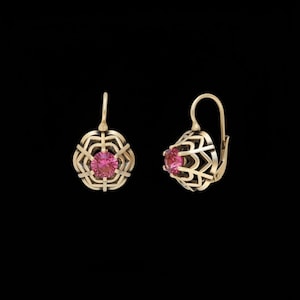 May include: A pair of gold-colored earrings with a pink gemstone center. The earrings have a filigree design around the gemstone and a hook closure. The background is black.