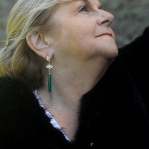 May include: A woman wearing a black fur coat with a pair of green gemstone dangle earrings. The earrings have a gold setting and a fan-shaped design.