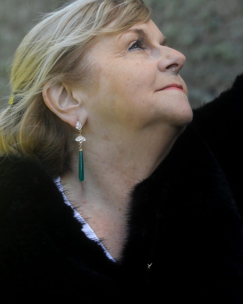 May include: A woman wearing a black fur coat with a pair of green gemstone dangle earrings. The earrings have a gold setting and a fan-shaped design.