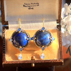 May include: Ornate gold earrings featuring large, round, deep blue lapis lazuli stones. The earrings have smaller blue and purple accent stones and a teardrop pendant. The earrings are presented in a vintage brown jewellery box.
