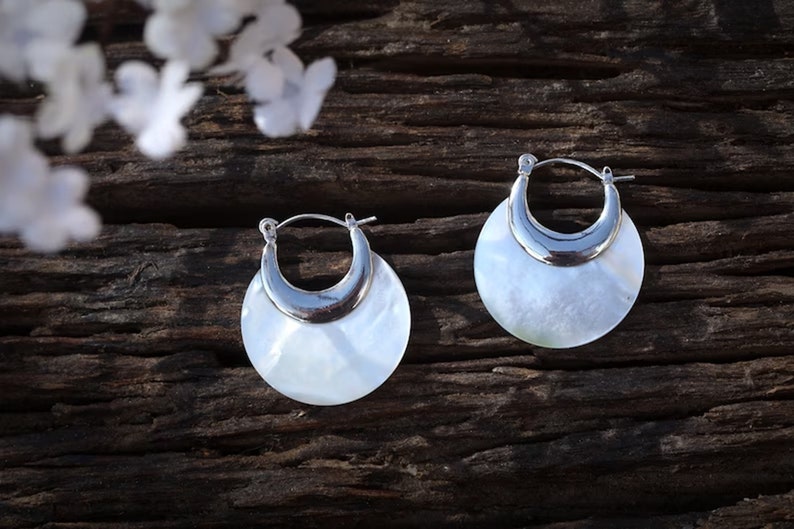 May include: A pair of silver hoop earrings with a white mother-of-pearl inlay. The earrings are round and have a crescent moon shape.