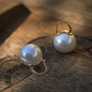 May include: A pair of gold hoop earrings with large white pearl accents. The earrings are on a wooden surface.