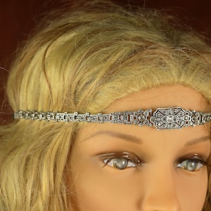 May include: A silver headband with a decorative, geometric design featuring clear rhinestones. The headband is worn on a mannequin with blonde hair.