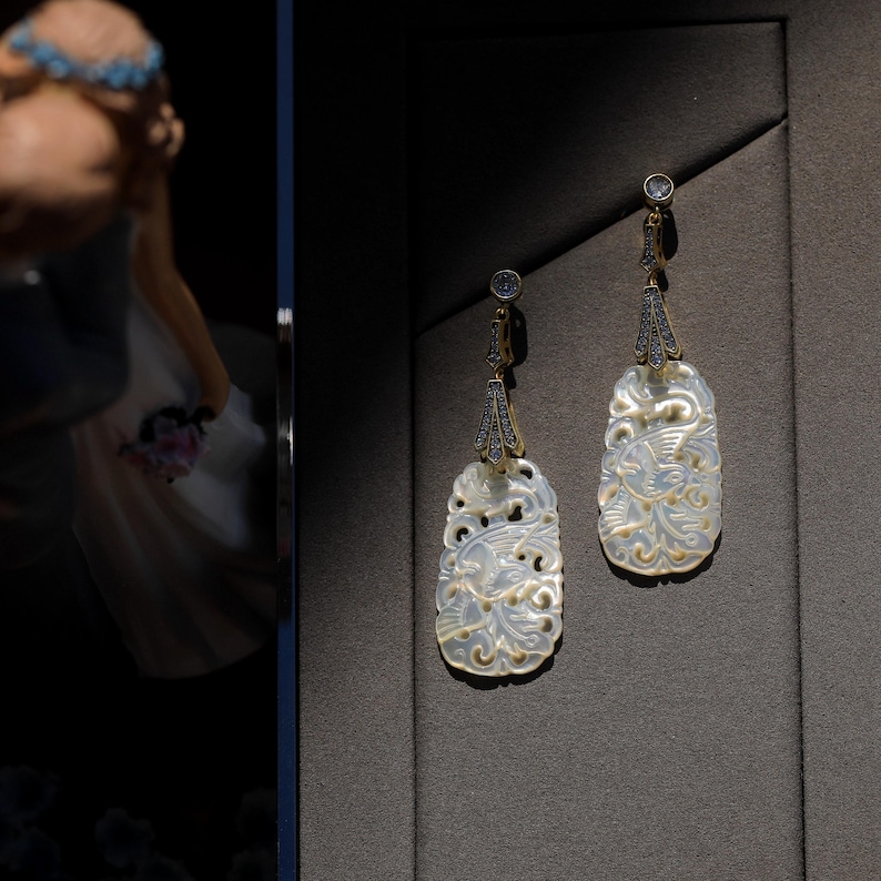 May include: A pair of white, intricately carved earrings with gold accents and clear stones. The earrings are oval shaped and feature a floral design.