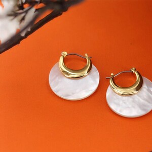 May include: A pair of gold hoop earrings with white mother-of-pearl accents. The earrings are round and have a crescent-shaped gold piece on top.