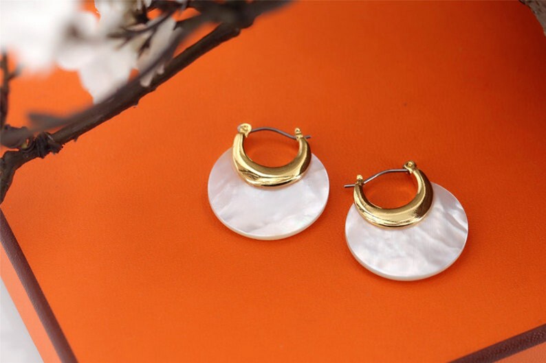 May include: A pair of gold hoop earrings with white mother-of-pearl accents. The earrings are round and have a crescent-shaped gold piece on top.