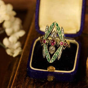 Pode incluir: Um anel de ouro com um design de lib&eacute;lula. As asas da lib&eacute;lula s&atilde;o de esmalte verde com diamantes brancos e o corpo &eacute; adornado com rubis vermelhos. O anel est&aacute; em uma caixa de anel de veludo azul.