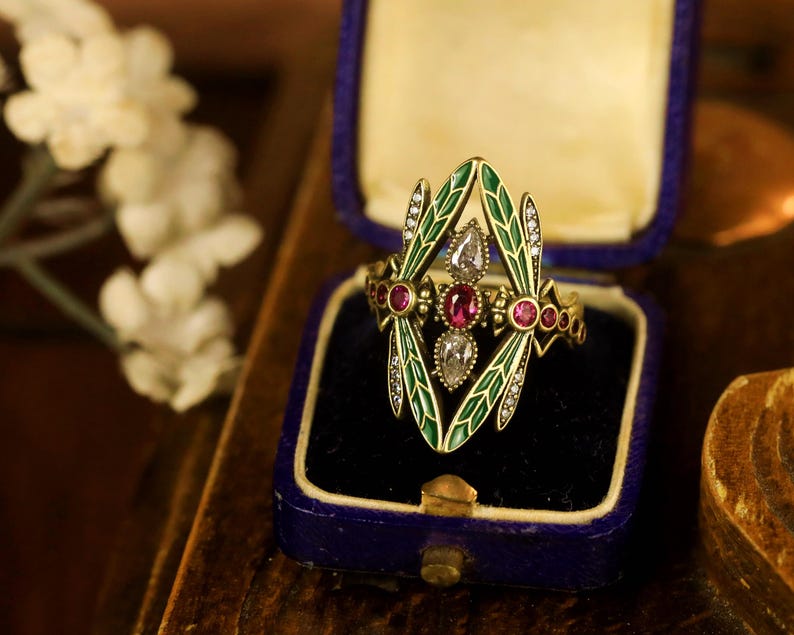 Pode incluir: Um anel de ouro com um design de lib&eacute;lula. As asas da lib&eacute;lula s&atilde;o de esmalte verde com diamantes brancos e o corpo &eacute; adornado com rubis vermelhos. O anel est&aacute; em uma caixa de anel de veludo azul.