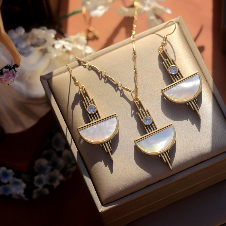 May include: A gold-toned jewelry set consisting of a necklace and two matching drop earrings. Each piece features a semi-circular white mother-of-pearl inlay, a small clear round stone, and vertical metal bars arranged in a geometric pattern.