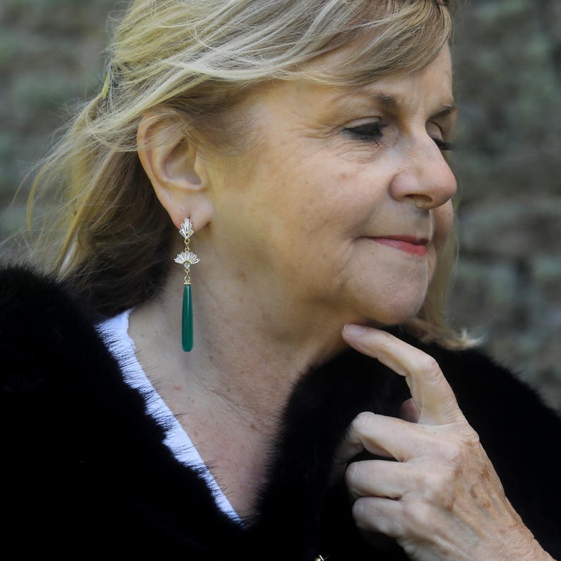 May include: A woman wearing a black fur coat with a pair of green and gold dangle earrings. The earrings have a teardrop-shaped green stone and a gold filigree design.