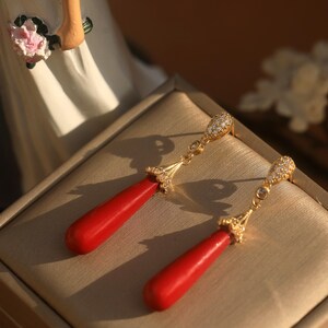 May include: A pair of gold-colored drop earrings with red teardrop-shaped pendants. The earrings have a decorative top with small, clear stones. The earrings are displayed on a beige surface.