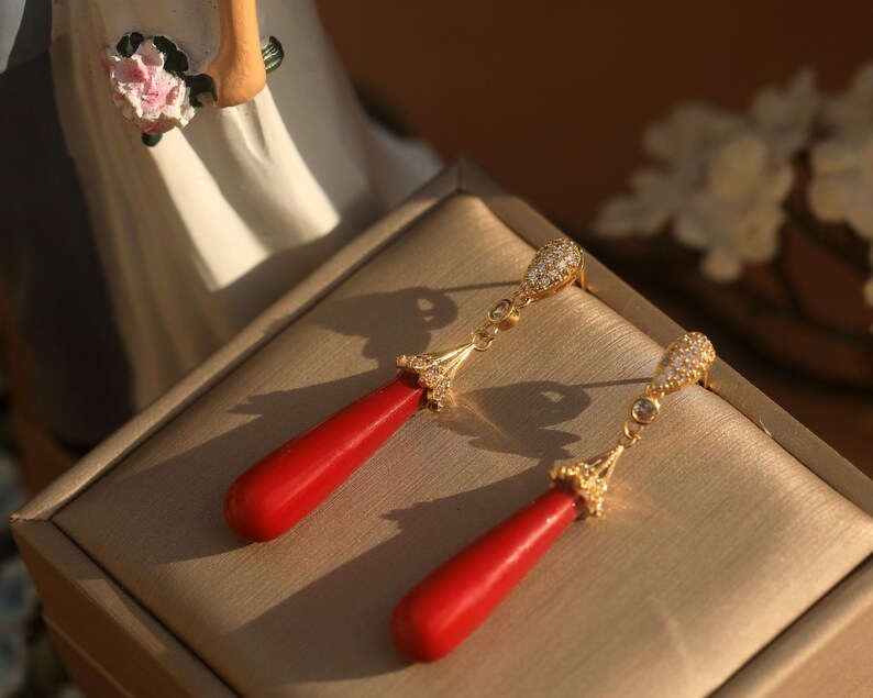 May include: A pair of gold-colored drop earrings with red teardrop-shaped pendants. The earrings have a decorative top with small, clear stones. The earrings are displayed on a beige surface.
