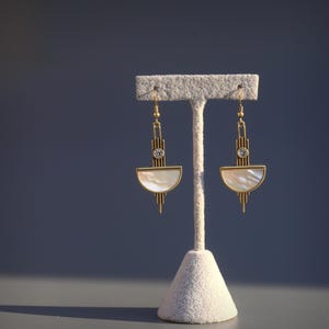 May include: A pair of gold-toned drop earrings featuring a vertical bar design with a small clear round stone at the center and a semi-circular white iridescent shell inlay at the base, suspended from thin gold-toned metal hooks.