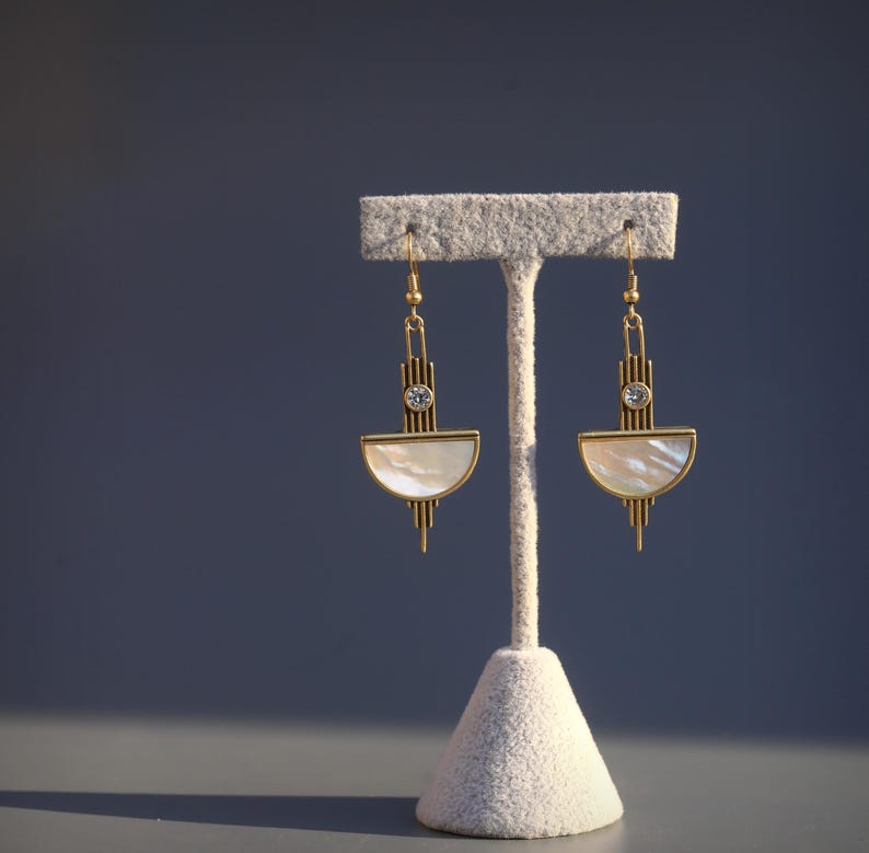 May include: A pair of gold-toned drop earrings featuring a vertical bar design with a small clear round stone at the center and a semi-circular white iridescent shell inlay at the base, suspended from thin gold-toned metal hooks.