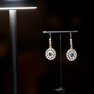 May include: A pair of gold earrings with a blue gemstone center surrounded by a circle of white rhinestones.