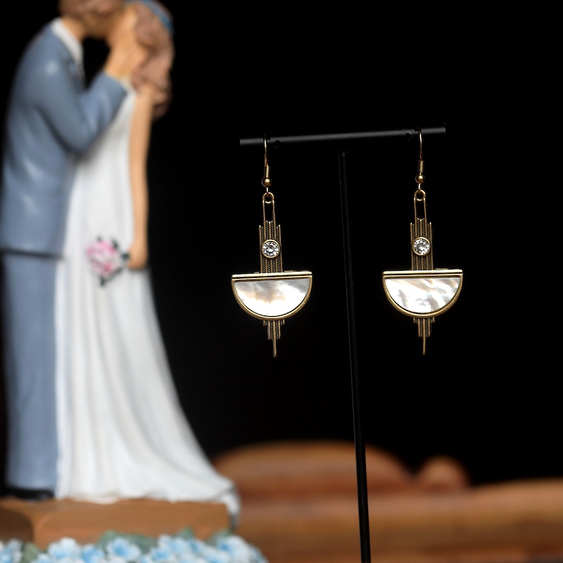 May include: A pair of gold-toned drop earrings featuring a semi-circular mother-of-pearl inlay set within a geometric frame. Each earring includes a small clear crystal accent positioned above the shell piece, suspended from a simple hook wire.