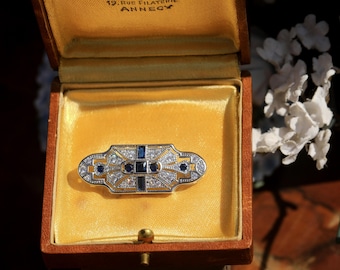 May include: An antique brooch in a wooden box. The brooch is rectangular with a gold-colored frame, featuring clear and blue gemstones. The box has a yellow satin lining. The brooch is a vintage jewelry item.