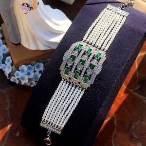Può includere: Un elegante bracciale con più fili di piccole perle bianche. Il punto focale è un elemento centrale rettangolare e ornato con dettagli in cristallo verde smeraldo e trasparente, incastonato in metallo argentato. Il bracciale è esposto su una superficie blu scuro.