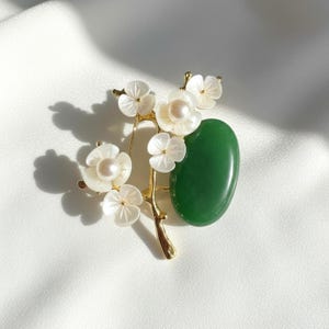 May include: A gold-toned brooch featuring a floral design with white mother-of-pearl flowers and a central pearl. A polished green stone is set next to the flowers. The brooch is set on a white background.