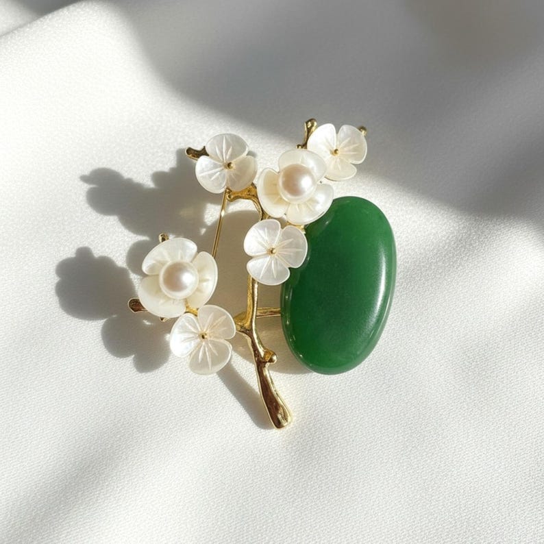 May include: A gold-toned brooch featuring a floral design with white mother-of-pearl flowers and a central pearl. A polished green stone is set next to the flowers. The brooch is set on a white background.