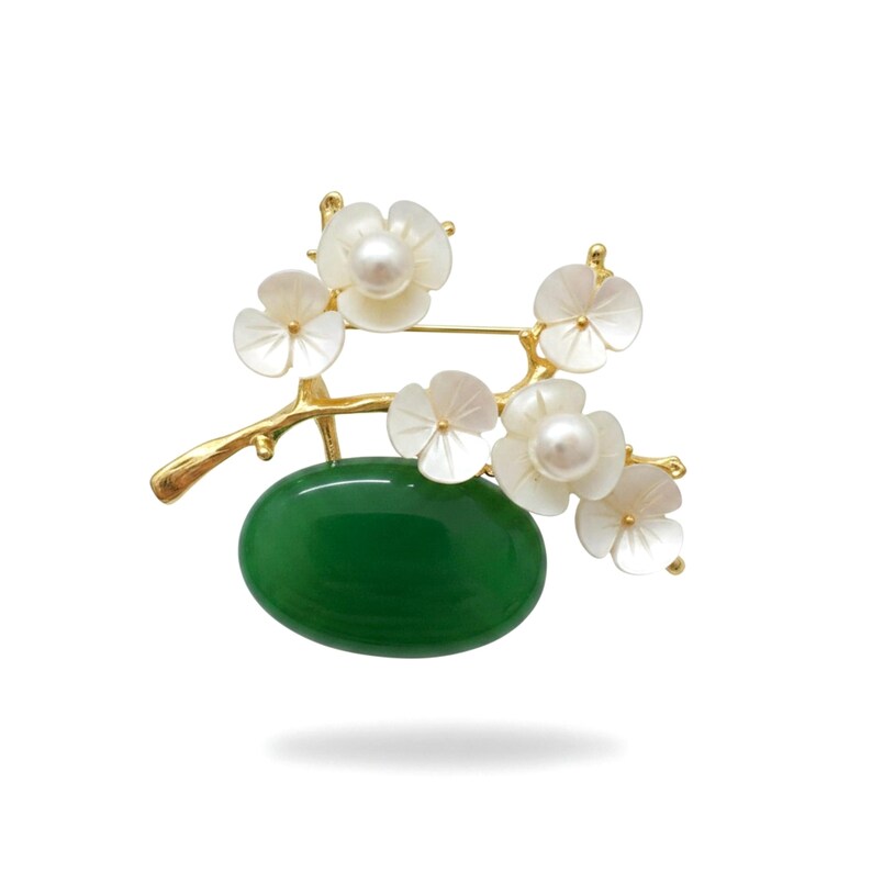 May include: A gold-toned brooch featuring a branch design with white flower accents and a central green oval stone. The flowers have pearl centers. The brooch is set against a white background.