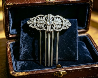 May include: A silver-toned hair comb featuring an intricate openwork design adorned with numerous small clear crystals and a single oval-shaped central stone. The decorative top section has a symmetrical pattern with curved lines and fan-like motifs.