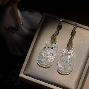 May include: A pair of white mother of pearl earrings with intricate floral designs and gold accents. The earrings are dangling and have a small clear crystal at the top.