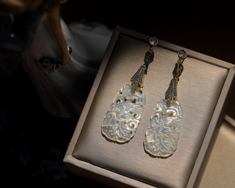 May include: A pair of white mother of pearl earrings with intricate floral designs and gold accents. The earrings are dangling and have a small clear crystal at the top.